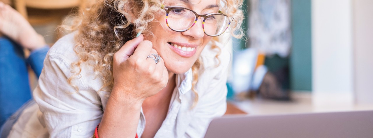 Μια γυναίκα γύρω στα 50 με κατσαρά ξανθά μαλλιά και γυαλιά κοιτάει στο laptop της. Είναι ξαπλωμένη φοράει άσπρο πουκάμισο και τζιν μπλε και εμείς βλέπουμε