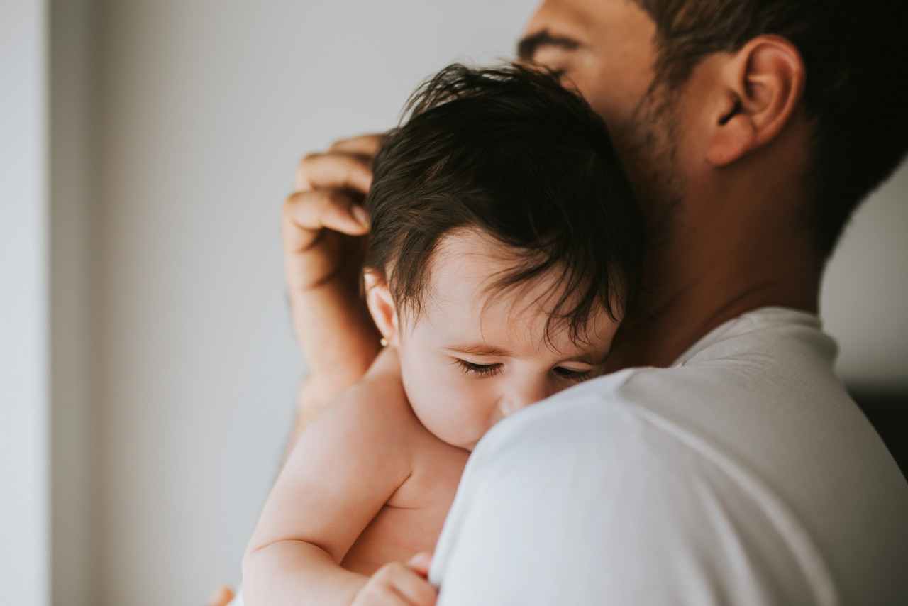 Ένας πατέρας κρατά το μωρό του στον ώμο του. Ο άντρας είναι μελαχρινός και δεν τον βλέπουμε καθαρά γιατί το μωρό που είναι γυμνό είναι στραμένο στην κάμερα. Του χαϊδευει το κεφάλι απαλά.